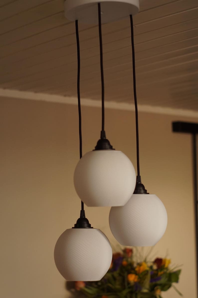 Pu&ograve; includere: Tre lampade a sospensione con paralumi a globo bianchi e cavi neri pendono da un apparecchio a soffitto bianco. I paralumi hanno un motivo strutturato e scanalato. Le luci sono sospese sopra un tavolo con una composizione floreale.