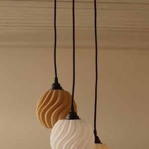 Pu&ograve; includere: Tre lampade a sospensione con supporto a soffitto bianco e cavi neri. Gli apparecchi di illuminazione hanno un design strutturato e vorticoso nei toni del giallo e del bianco. Le luci sono appese su uno sfondo neutro.