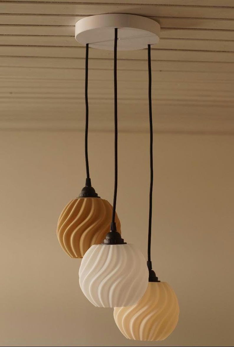 Pu&ograve; includere: Tre lampade a sospensione con supporto a soffitto bianco e cavi neri. Gli apparecchi di illuminazione hanno un design strutturato e vorticoso nei toni del giallo e del bianco. Le luci sono appese su uno sfondo neutro.