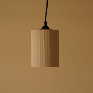 Pu&ograve; includere: Un lampadario cilindrico con un paralume color crema a coste verticali. La luce &egrave; sospesa da un cavo e un apparecchio neri su uno sfondo neutro. Il design del paralume aggiunge consistenza al design minimalista.