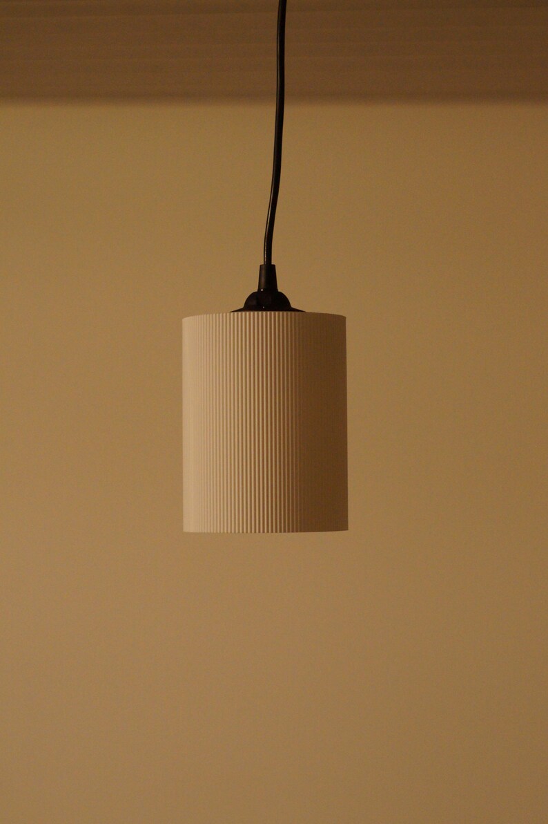 Pu&ograve; includere: Un lampadario cilindrico con un paralume color crema a coste verticali. La luce &egrave; sospesa da un cavo e un apparecchio neri su uno sfondo neutro. Il design del paralume aggiunge consistenza al design minimalista.