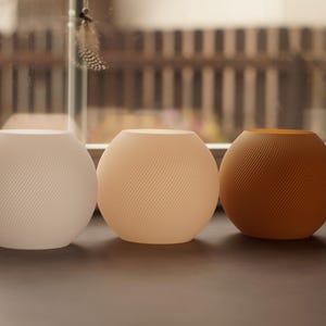 Pu&ograve; includere: Tre oggetti decorativi sferici in fila. Gli oggetti sono nei toni del bianco, pesca e marrone. Ognuno ha un design a spirale strutturato. Sono su una superficie scura, con una finestra e vista esterna sullo sfondo.