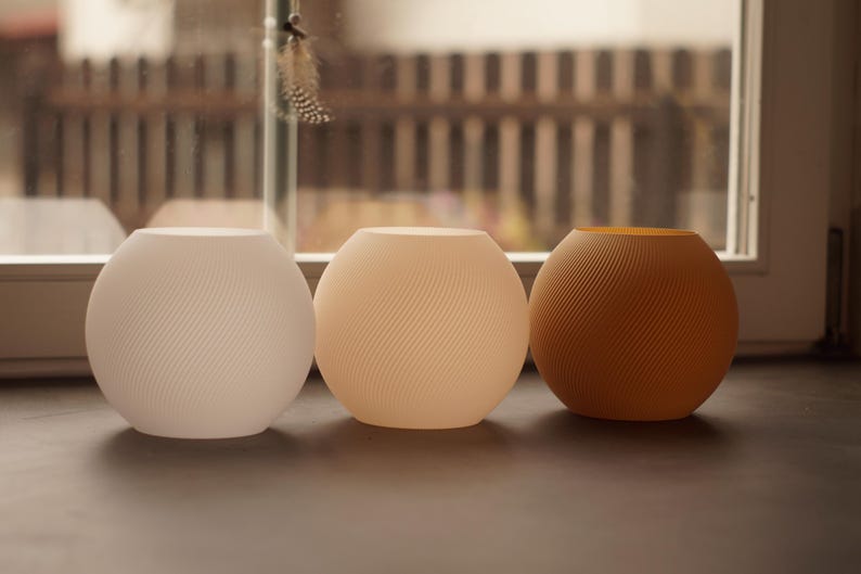 Pu&ograve; includere: Tre oggetti decorativi sferici in fila. Gli oggetti sono nei toni del bianco, pesca e marrone. Ognuno ha un design a spirale strutturato. Sono su una superficie scura, con una finestra e vista esterna sullo sfondo.