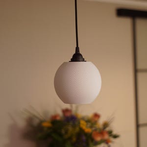 Pu&ograve; includere: Una lampada a sospensione sferica bianca con un design a spirale strutturato. La lampada &egrave; appesa a un cavo e un apparecchio neri. Un bouquet sfocato di fiori in un vaso bianco &egrave; in primo piano.