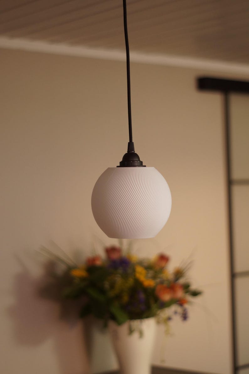 Pu&ograve; includere: Una lampada a sospensione sferica bianca con un design a spirale strutturato. La lampada &egrave; appesa a un cavo e un apparecchio neri. Un bouquet sfocato di fiori in un vaso bianco &egrave; in primo piano.