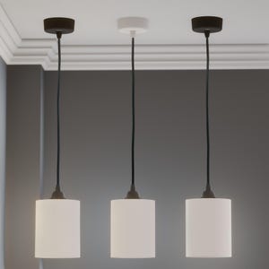 Pu&ograve; includere: Tre lampade a sospensione con paralumi cilindrici bianchi e cavi neri. I supporti a soffitto sono neri e bianchi. I paralumi hanno linee verticali. Le lampade sono sospese contro un muro grigio.