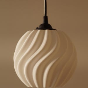 Pu&ograve; includere: Una lampada a sospensione sferica bianca con un design ondulato e strutturato. Pende da un cavo nero e da un supporto a soffitto nero. La lampada &egrave; di design moderno.