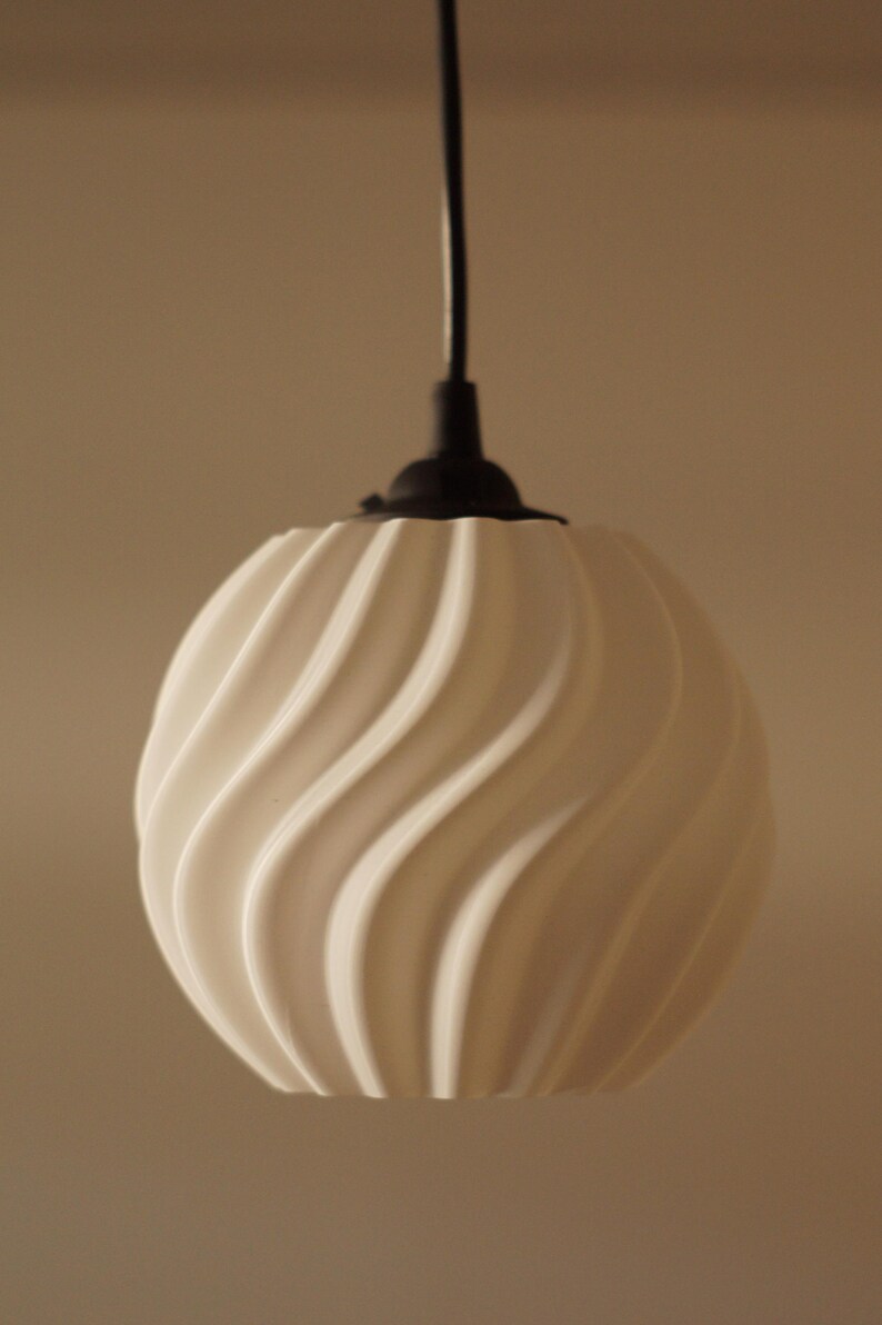 Pu&ograve; includere: Una lampada a sospensione sferica bianca con un design ondulato e strutturato. Pende da un cavo nero e da un supporto a soffitto nero. La lampada &egrave; di design moderno.