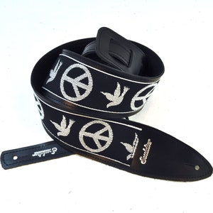 以下が含まれることがあります： 白い平和マークと鳩が刺繍された黒いギターストラップ。ストラップは革製でバックルが付いています。