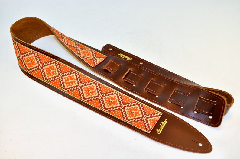 K&ouml;nnte beinhalten: Braunes Ledergitarrengurt mit einem orangefarbenen und cremefarbenen Stoff-Einsatz. Das Gurt hat eine Schnalle und ist in der L&auml;nge verstellbar. Das Gurt ist mit "Souldier" in goldenen Buchstaben beschriftet.