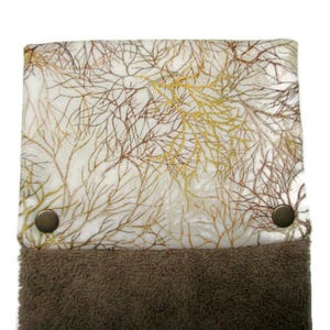 May include: A decorative kitchen towel featuring a white backdrop adorned with a pattern of brown and gold branches. The towel has a brown, plush bottom and is secured with two bronze-coloured press studs.