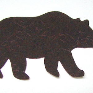May include: A black bear silhouette cut out of a dark brown material with a textured pattern. The bear is facing right and walking.