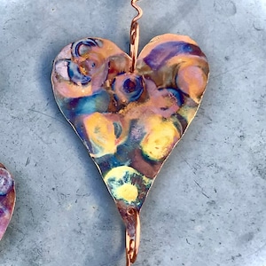 May include: A copper heart with a swirling, iridescent pattern hangs from a copper wire. The heart is attached to a red glass bead.