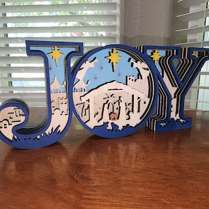 May include: A wooden JOY sign with a blue background and a nativity scene cutout. The scene includes a stable, a star, and three wise men.