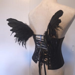 May include: A black corset featuring a lace-up back with silver-toned metal grommets and black ribbon ties. Attached to the upper back of the corset are two wings constructed from black feathers, extending outward from the shoulder blade area.