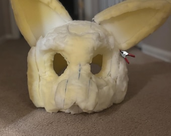 Może przedstawiać: Częściowo zbudowana baza głowy fursuit w kolorze jasnożółtym i białym. Głowa ma duże, wyprostowane uszy i otwory na oczy. Niebieskie linie wyznaczają wzór. Dołączony jest mały zacisk z czerwonymi i czarnymi przewodami.