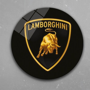 May include: A round, black emblem featuring the Lamborghini logo. The logo includes a gold bull inside a gold-bordered shield, with "LAMBORGHINI" in gold lettering above. The emblem is set against a gray background.