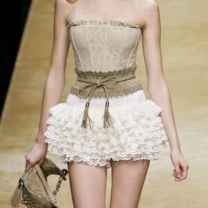 May include: A beige strapless corset top with a lace design, paired with a white ruffled mini skirt. A brown belt with tassels cinches the waist. The model is carrying a beige handbag.