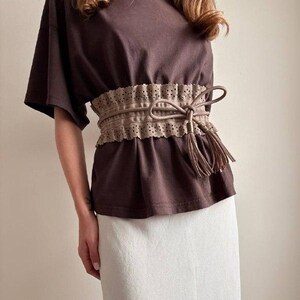 May include: A brown t-shirt paired with a white skirt. A tan belt with a bow and tassels cinches the waist. The belt has a decorative, perforated edge. The outfit is simple and elegant.
