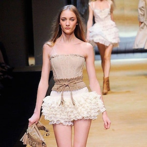 May include: A model on a runway wearing a beige corset top, a white ruffled skirt, and brown boots. A brown belt with tassels cinches the waist. The model carries a small, beige handbag with a chain.