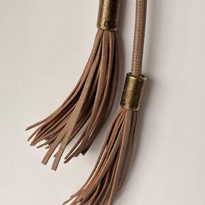 May include: Close-up of two brown leather tassels with gold-colored metal caps. The caps have the letters "D.G." engraved on them. The tassels are hanging against a white background.