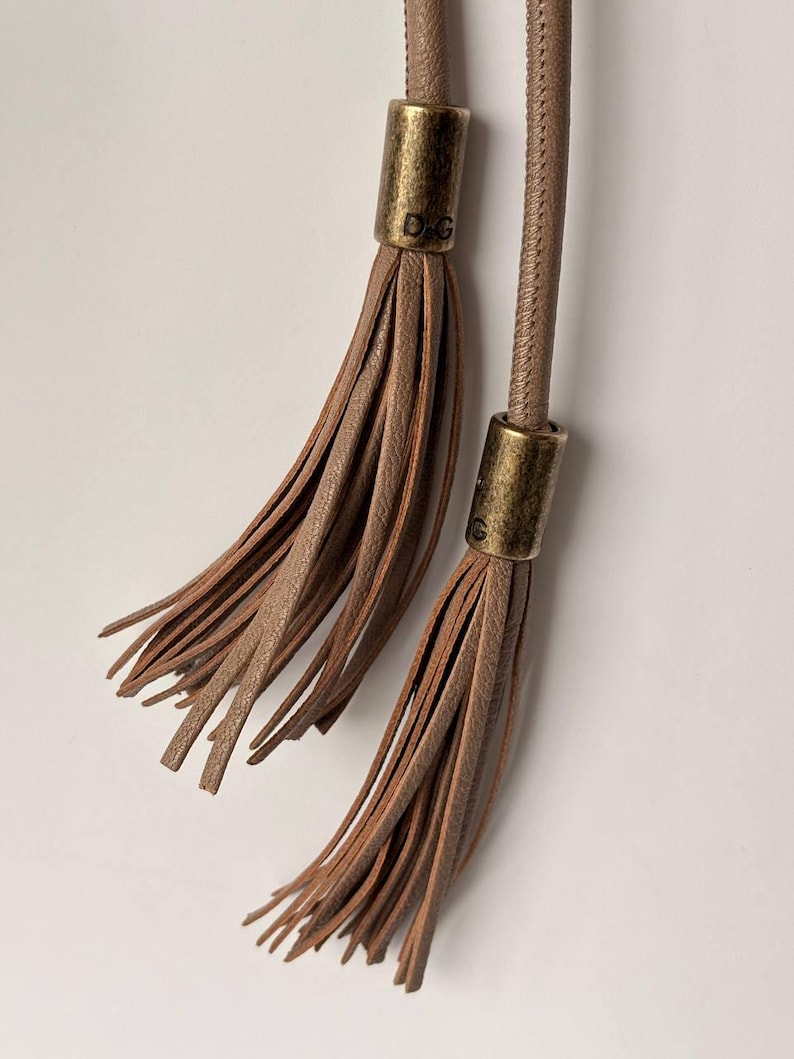 May include: Close-up of two brown leather tassels with gold-colored metal caps. The caps have the letters "D.G." engraved on them. The tassels are hanging against a white background.