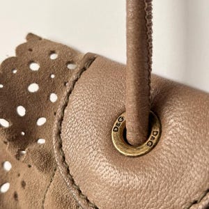May include: Close-up of a tan handbag with a handle. The bag features a laser-cut design on the suede-like material. The handle is a twisted, stitched leather. A gold-colored metal ring with the letters "D&G" is visible.