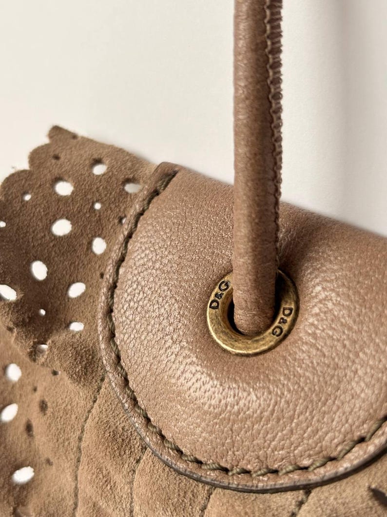 May include: Close-up of a tan handbag with a handle. The bag features a laser-cut design on the suede-like material. The handle is a twisted, stitched leather. A gold-colored metal ring with the letters "D&G" is visible.
