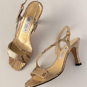 Könnte beinhalten: Ein Paar goldfarbene Sandalen mit offenem Zehenbereich und hohem Absatz mit Schlangenmuster. Die Sandalen haben dünne Riemen, einen Schnallenverschluss und einen kleinen Absatz. Der Markenname "LANVIN PARIS" ist auf der Innensohle sichtbar.