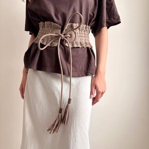 May include: A beige, wide waist belt with a decorative, perforated design. The belt has a tie closure with long, dangling cords and tassel ends. The belt is worn over a brown t-shirt and a white skirt.