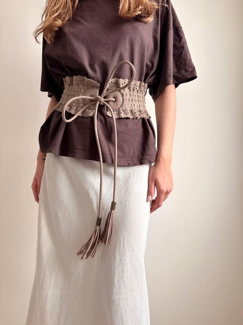 May include: A beige, wide waist belt with a decorative, perforated design. The belt has a tie closure with long, dangling cords and tassel ends. The belt is worn over a brown t-shirt and a white skirt.