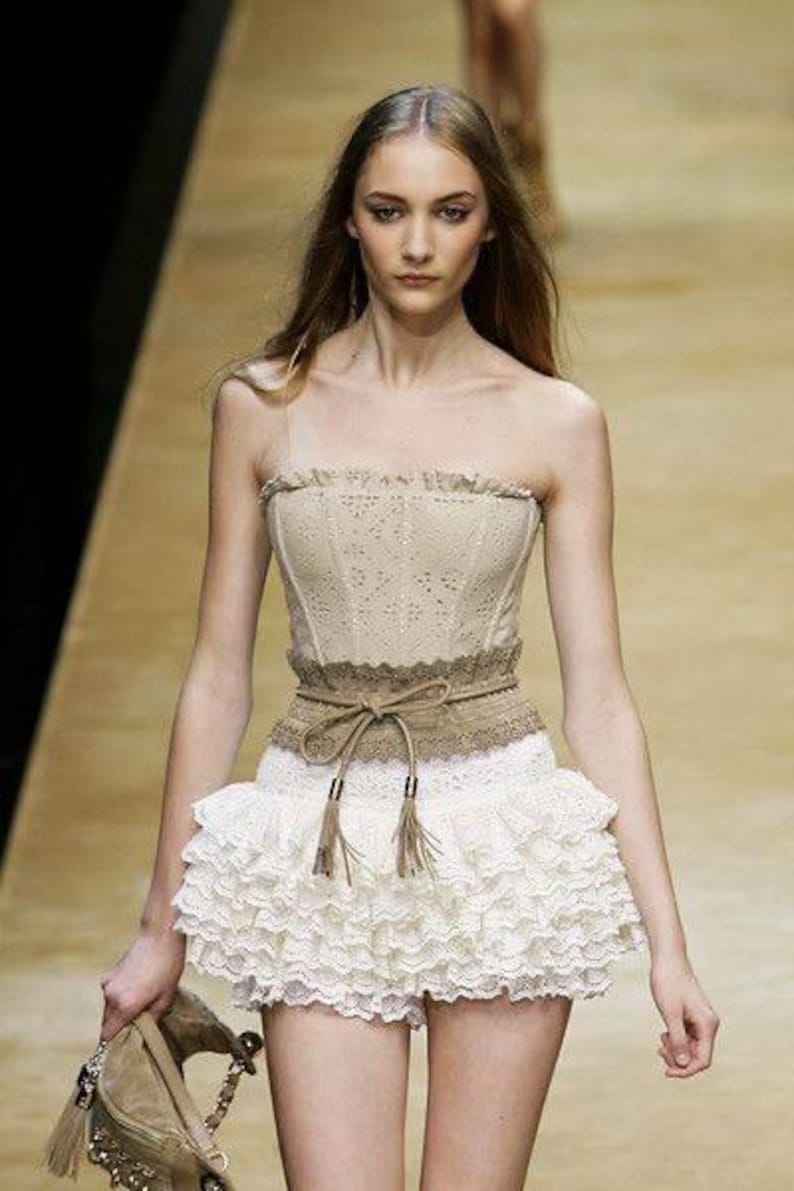 May include: A beige strapless corset top with a lace design, paired with a white ruffled mini skirt. A brown belt with tassels cinches the waist. The model is carrying a beige handbag.