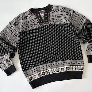 Puede incluir: Un suéter de manga larga en blanco y negro con un patrón geométrico detallado y copos de nieve en el pecho, los hombros y el dobladillo. El cuello incluye una tapeta corta con tres botones metálicos y un borde oscuro con sutiles toques rojos.