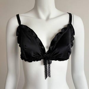 Puede incluir: Sujetador de satén negro con bordes con volantes y delicados detalles de encaje. El sujetador tiene tirantes ajustables y un pequeño lazo en el centro. Se muestra en un maniquí blanco.