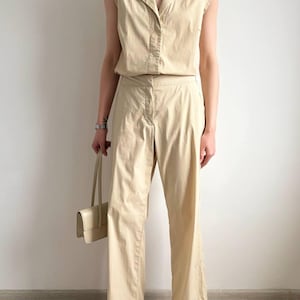 May include: A beige two-piece outfit. It includes a collared, sleeveless top and matching trousers. The outfit is accessorized with a small, light-coloured handbag and white sandals. The trousers have a straight leg design. The outfit is made of a light-coloured fabric.