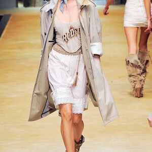 May include: A model on a runway wears a beige trench coat over a cream-colored top with the word "RODEO" and white shorts with lace trim. She is wearing sunglasses and brown cowboy boots. Another model is in the background.