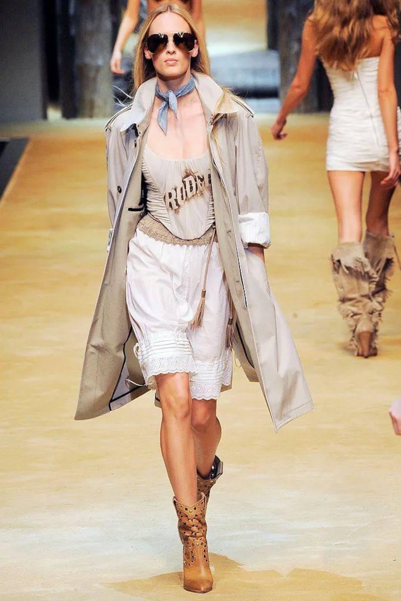 May include: A model on a runway wears a beige trench coat over a cream-colored top with the word "RODEO" and white shorts with lace trim. She is wearing sunglasses and brown cowboy boots. Another model is in the background.