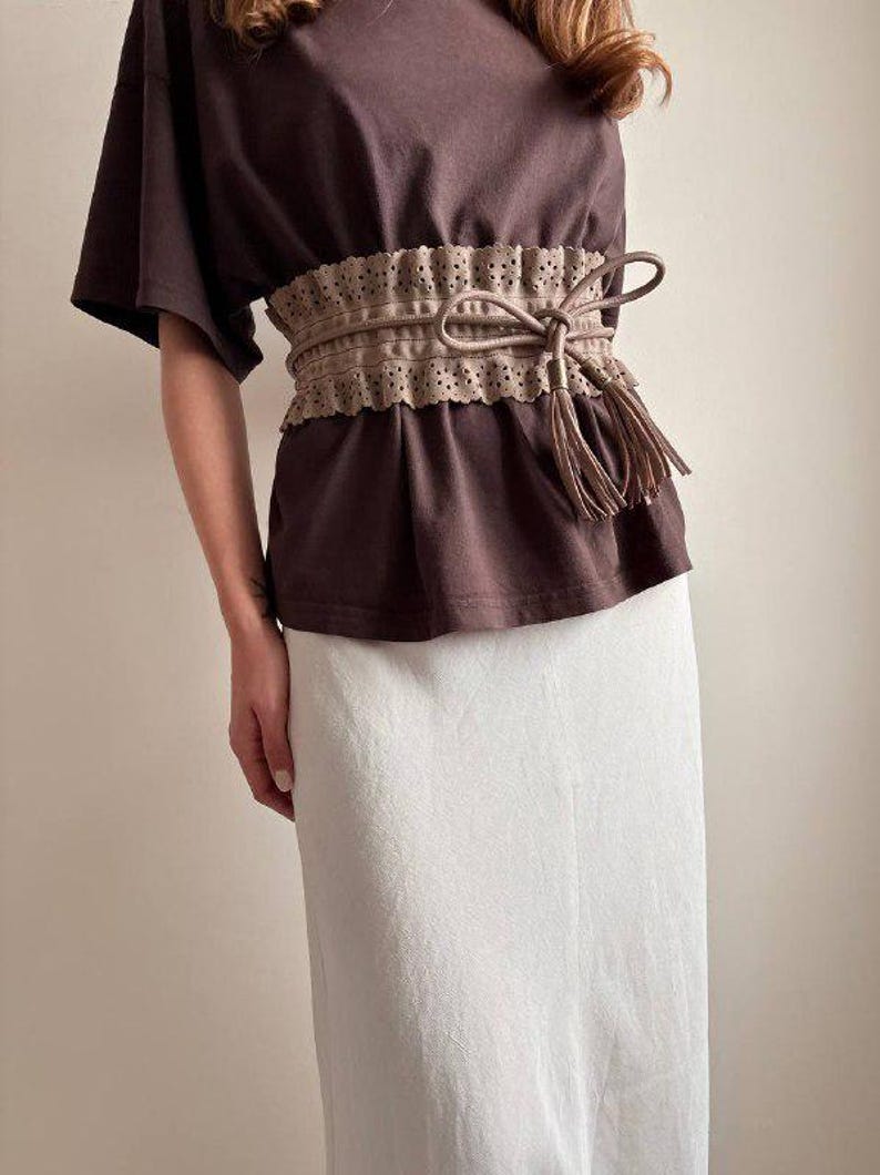 May include: A brown t-shirt paired with a white skirt. A tan belt with a bow and tassels cinches the waist. The belt has a decorative, perforated edge. The outfit is simple and elegant.
