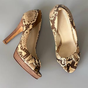 Op de afbeelding: Een paar pumps met hoge hakken en een slangenleerpatroon in beige en donkerbruine tinten. De schoenen hebben een houten plateauzool en hak, met een goudkleurig metalen bitdetail aan de voorzijde. De binnenvoering is lichtbeige van kleur.