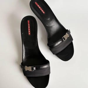 Può includere: Un paio di sandali neri Prada. I sandali presentano un piccolo tacco, un cinturino nero con fibbia argentata e il logo Prada in rosso e bianco. Sono esposti su sfondo bianco.