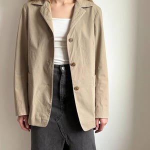May include: A tan blazer with three brown buttons and two front pockets. The blazer is worn over a white top and paired with a dark gray denim skirt. The outfit is simple and classic.