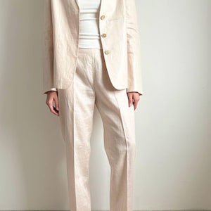 May include: A light peach-colored linen suit, consisting of a blazer and matching trousers. The blazer has three buttons and a notched lapel. The trousers are straight-legged. A white top is worn underneath the blazer. The outfit is paired with white sandals.