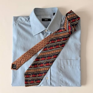 Könnte beinhalten: Hellblaues Hemd mit BOSS-Etikett und gemusterter Krawatte. Die Krawatte hat ein sich wiederholendes geometrisches Design in Braun und Gold mit roten floralen Akzenten auf marineblauem Hintergrund.