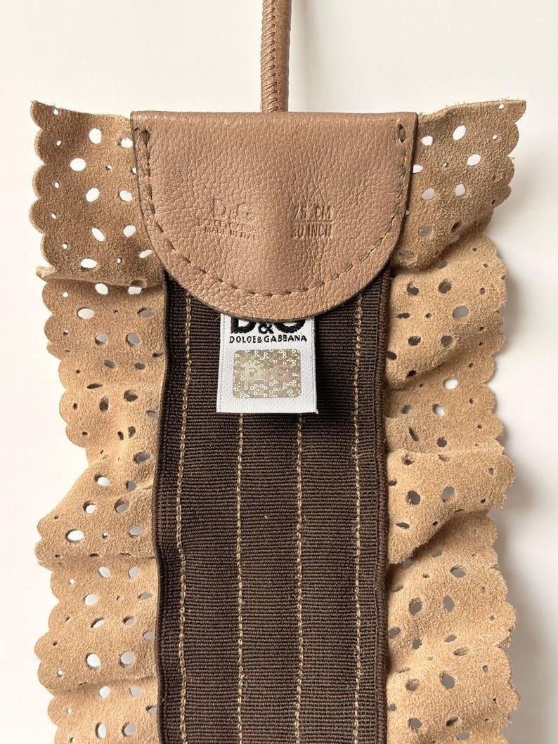 May include: A tan leather accessory with a perforated edge and a brown striped center. The accessory has a small leather flap with the brand name and measurements of 75 cm / 30 inch. A brand tag is visible.