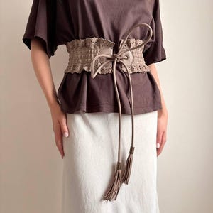 May include: A tan-colored, wide waist belt with a decorative, perforated design. The belt features long, dangling ties with tassel ends. The belt is worn over a brown top and a white skirt.
