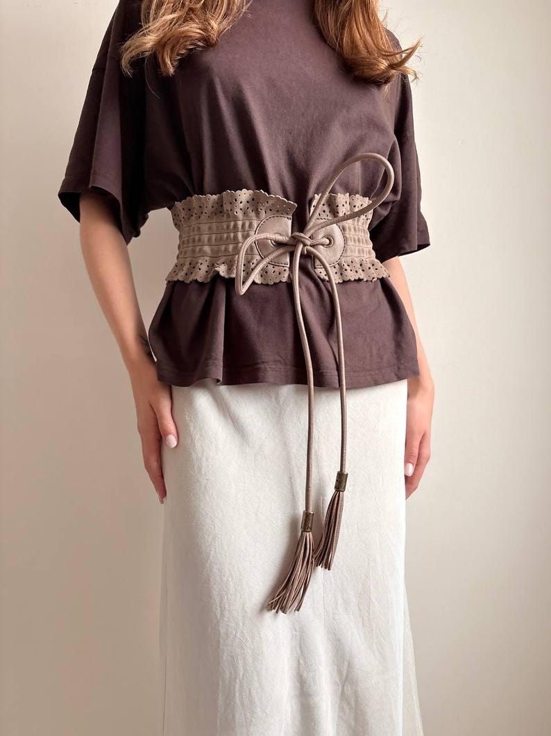 May include: A tan-colored, wide waist belt with a decorative, perforated design. The belt features long, dangling ties with tassel ends. The belt is worn over a brown top and a white skirt.