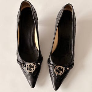 May include: A pair of black leather pumps with pointed toes. The shoes feature a silver-tone buckle with the Gucci logo and small decorative studs. The heels are approximately 3 inches high. The leather has a perforated design.