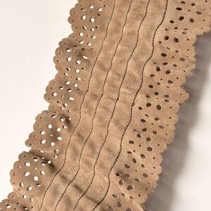 May include: Close-up of a tan leather belt with a scalloped edge and decorative perforations. The belt features vertical stitched lines and a soft, suede-like texture. The neutral color makes it versatile for various outfits.