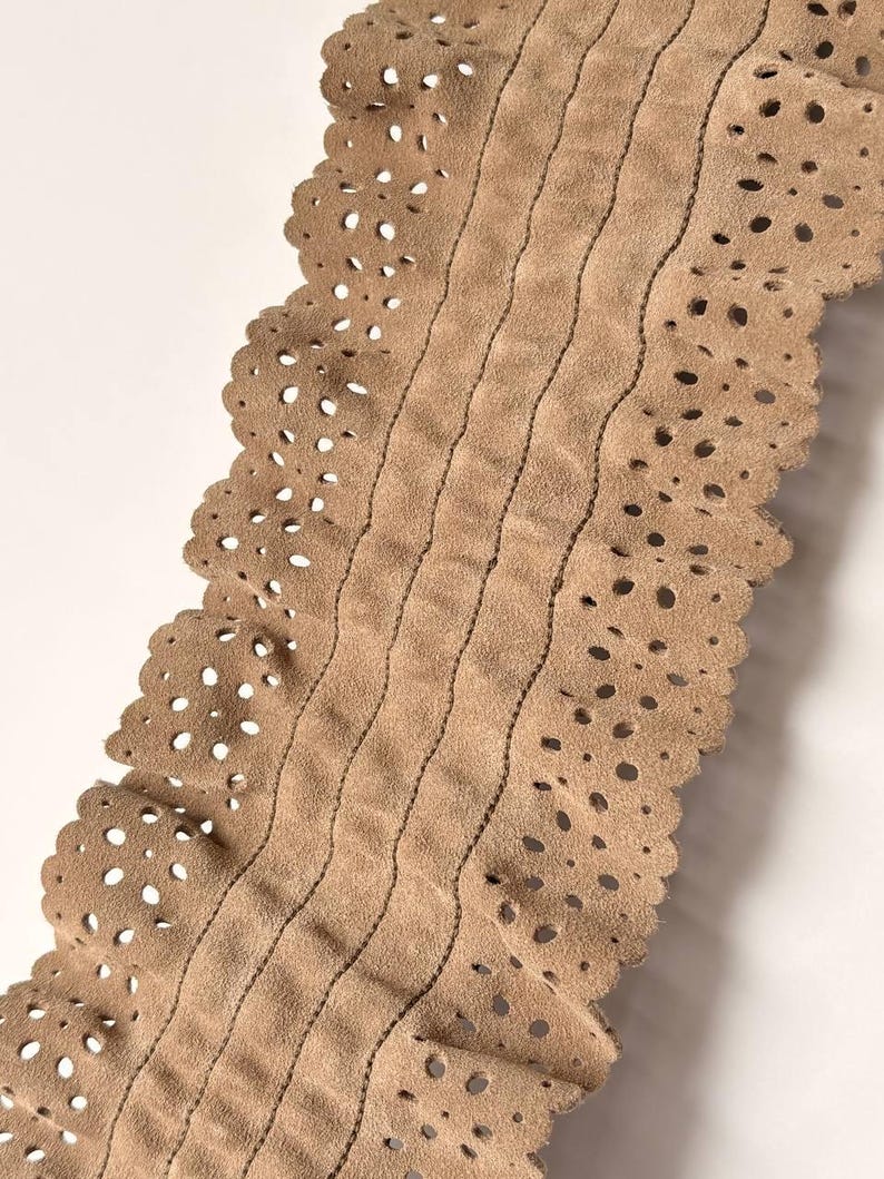 May include: Close-up of a tan leather belt with a scalloped edge and decorative perforations. The belt features vertical stitched lines and a soft, suede-like texture. The neutral color makes it versatile for various outfits.