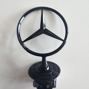 May include: Black Mercedes-Benz car emblem featuring a three-pointed star within a circular ring. The emblem is mounted on a black base with a silver screw.